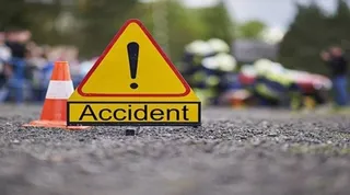 Un semn de avertizare triunghiular galben cu un punct exclamării și cuvântul Accident este plasat pe un drum lângă un con portocaliu.