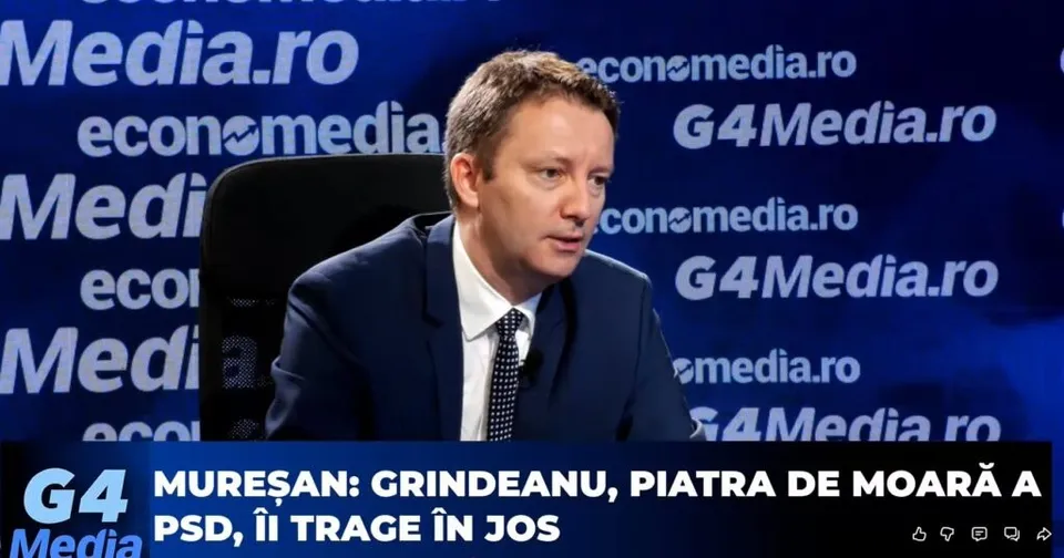 Un bărbat cu părul scurt, purtând sacou închis și cămașă albă cu cravată, stă în fața unui fundal albastru cu diverse logouri de site-uri de știri.