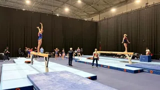 Mai mulți gimnaști execută mișcări pe aparate într-o sală de antrenament cu saltele albastre și albe și draperii negre pe fundal.