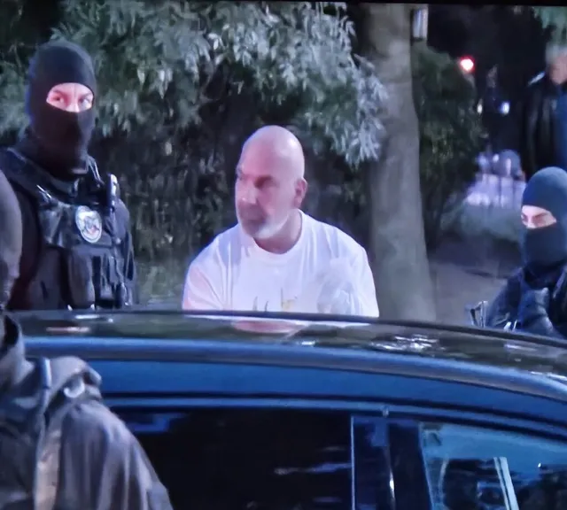 Un bărbat cu tricou alb stă lângă o mașină înconjurat de persoane purtând măști negre și echipamente tactice.