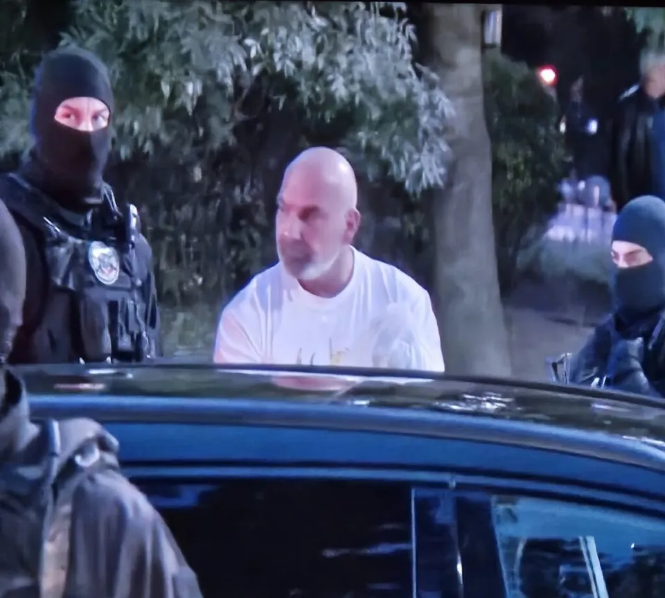 Un bărbat cu tricou alb stă lângă o mașină înconjurat de persoane purtând măști negre și echipamente tactice.