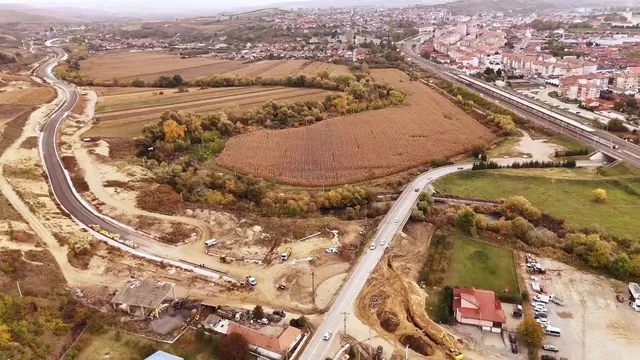 O vedere aeriană prezintă o porțiune de drum nou asfaltat, câmpuri agricole și o zonă urbană în fundal.