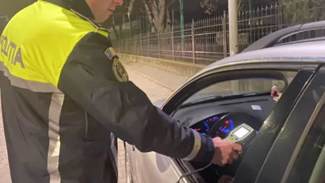 Un agent de poliție în uniformă portocalie și neagră folosește un aparat de testare în interiorul unui autoturism gri.