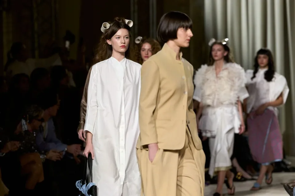 Patru modele merg pe o pistă de catwalk purtând haine de diverse culori și texturi în fața unui public așezat în umbră.