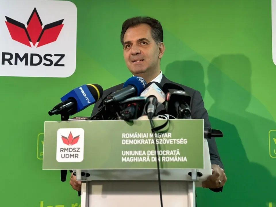 Un bărbat într-un costum închis la culoare stă în spatele unui podium pe care sunt amplasate mai multe microfoane în fața unui fundal verde.