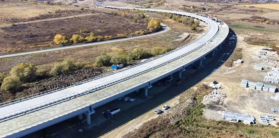 O vedere aeriană prezintă un viaduct lung și curbat cu parapeti albi, situat într-un peisaj de dealuri cu vegetație autumnală și materiale de construcție.