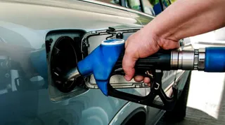 O mână ține un pistol de pompat combustibil albastru și negru introdus în orificiul de umplere al unui autovehicul gri.