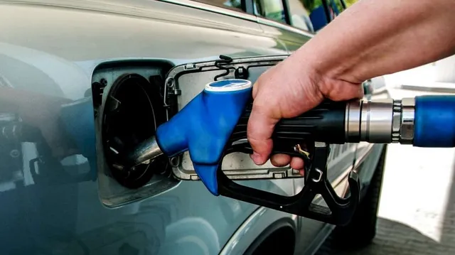 O mână ține un pistol de pompat combustibil albastru și negru introdus în orificiul de umplere al unui autovehicul gri.