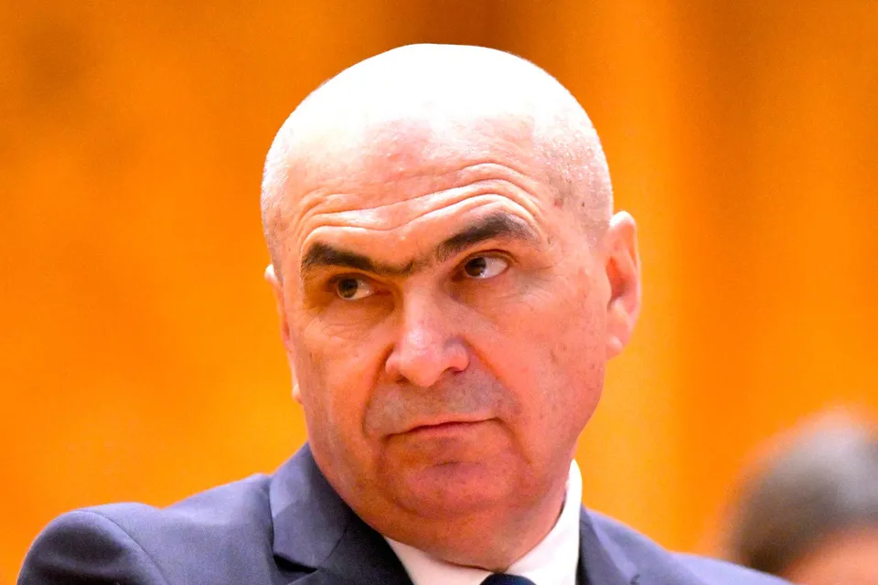 Un bărbat cu capul chel și sprâncene groase poartă un sacou închis la culoare și o cămașă albă.