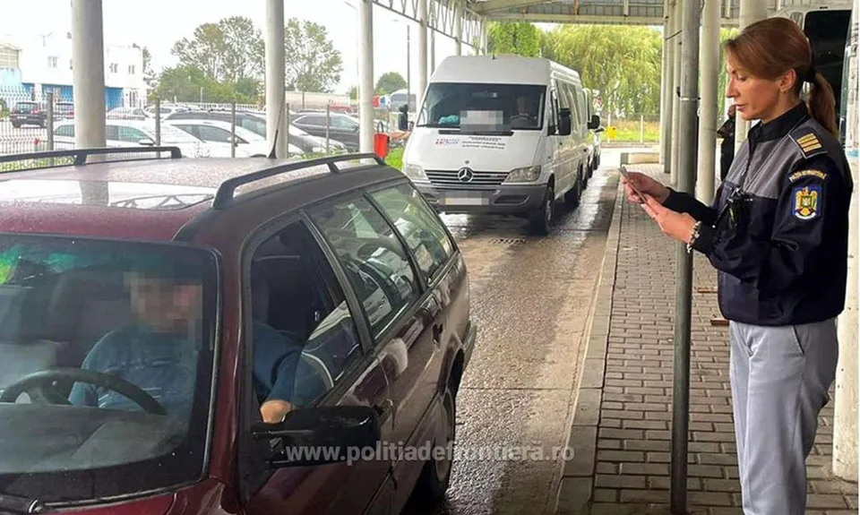 O polițistă în uniformă stă pe un trotuar lângă un van alb, în timp ce un autoturism maro trece pe lângă ea.
