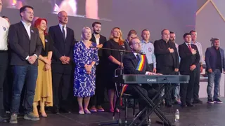 Un grup de persoane stă în picioare pe o scenă lângă un bărbat care este așezat la un instrument muzical de tip claviatură.