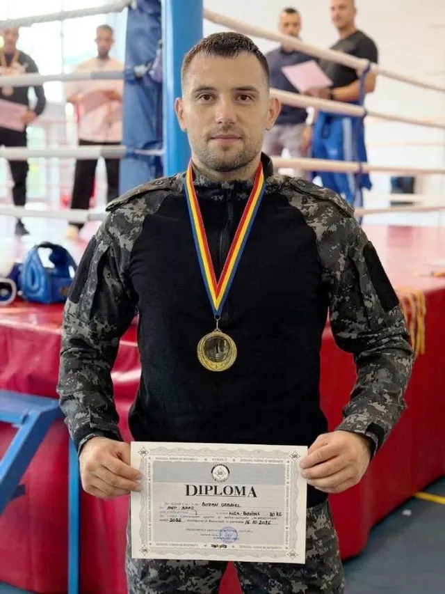 Un bărbat într-un costum militar gri și negru poartă o medalie aurie și ține un diplon în față într-un ring de sport.