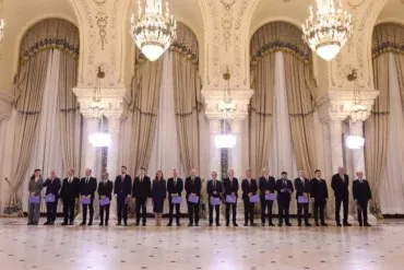 Un grup de oameni în costume oficiale stă aliniat într-o încăpere încărcată cu draperii mari și candelabre suspendate.