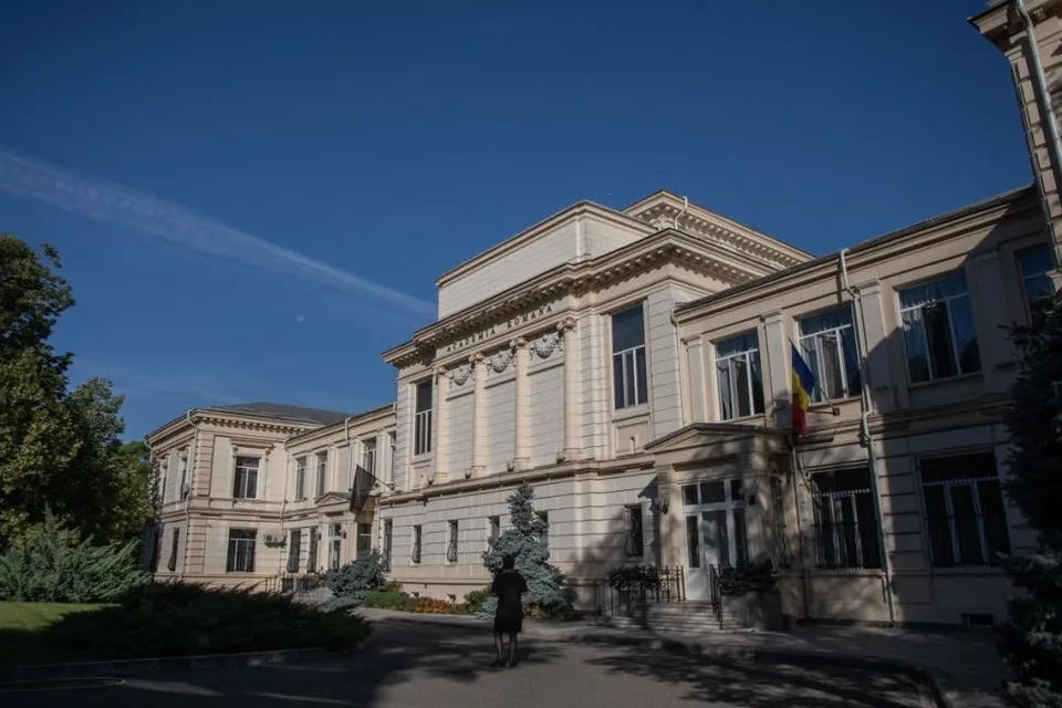 O persoană stă în fața unei clădiri mari, de culoare deschisă, sub un cer albastru.