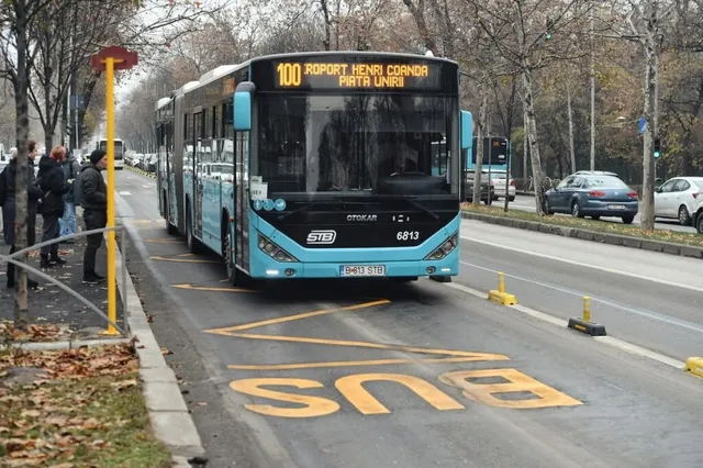 Un autobuz albastru cu numărul 100 circulă pe o bandă marcată cu textul BUS pe asfaltul unei străzi urbane.