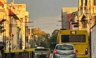 Un autobuz galben și o mașină gri circulă pe un drum urban flancat de clădiri și stâlpi de iluminat sub o lumină caldă.