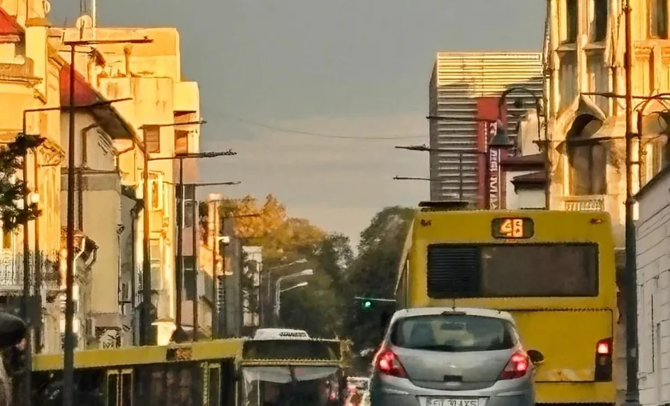 Un autobuz galben și o mașină gri circulă pe un drum urban flancat de clădiri și stâlpi de iluminat sub o lumină caldă.