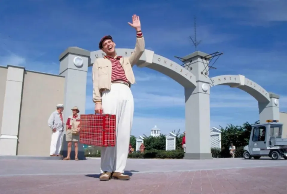Un bărbat cu pălărie, sacou bej și pantaloni albi stă în picioare salutând cu mâna, purtând un rucsac roșu în lateral.