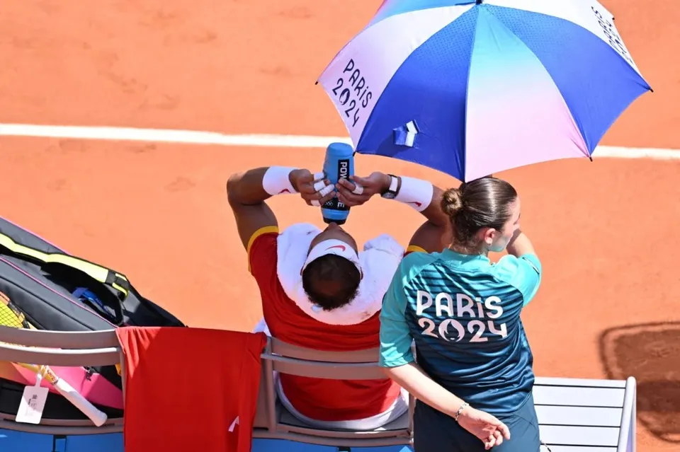 Un bărbat cu tricou roșu și bandană albă își toarnă apă dintr-o sticlă albastră pe cap, sub o umbrelă albastră și albă.