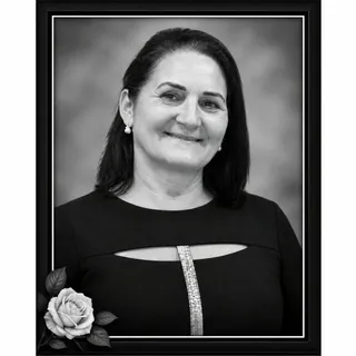 O femeie zâmbindă cu părul negru și haine negre este prezentată într-o fotografie de tip portret încadrată, lângă o petală de trandafir în colțul din stânga jos.