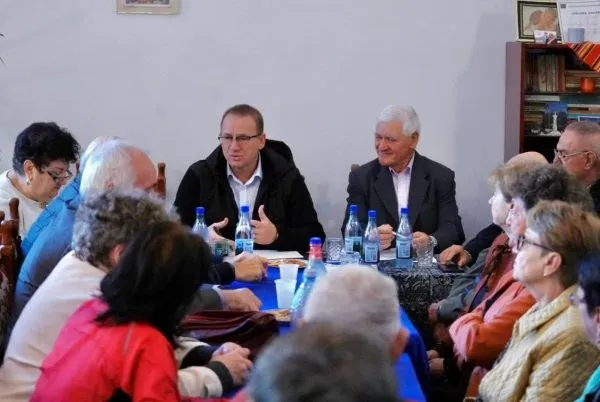 Un grup de persoane stă la o masă cu sticle de apă și pahare, în timp ce doi bărbați discută în partea de sus.