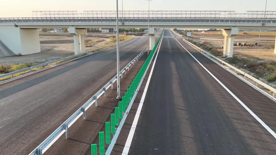 O autostradă cu asfalt negru dispune de linii albe pe margini, o barieră metalică și un gard verde central sub un pod de beton.
