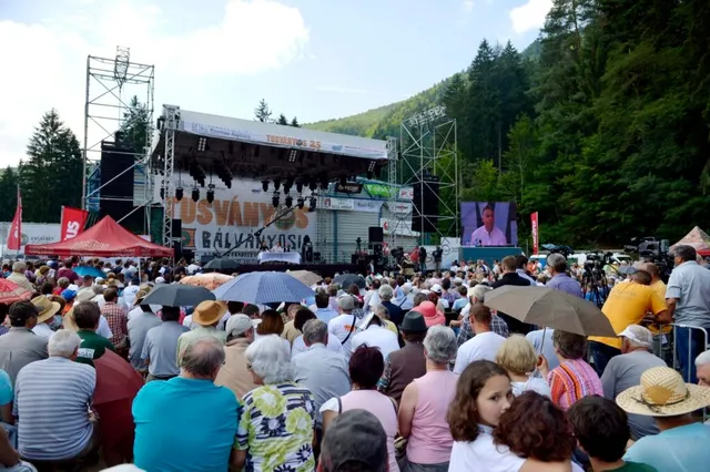 O mulțime de oameni stă cu spatele la cameră în fața unei scene de concerte cu panouri și ecrane mari sub un cer luminos.