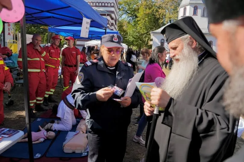 O femeie în uniformă neagră ține o broșură, în timp ce un bărbat cu barbă lungă și pălărie neagră stă în prim-plan.