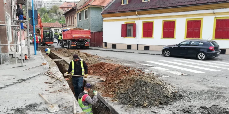 Oameni în veste reflectorizante lucrează la o șanț deschis pe un drum asfaltat lângă un camion roșu și clădiri.