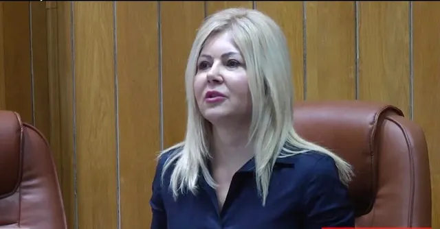 O femeie cu părul blond și o cămașă închisă la culoare stă în fața unui perete din panouri de lemn și a unor scaune de piele maro.