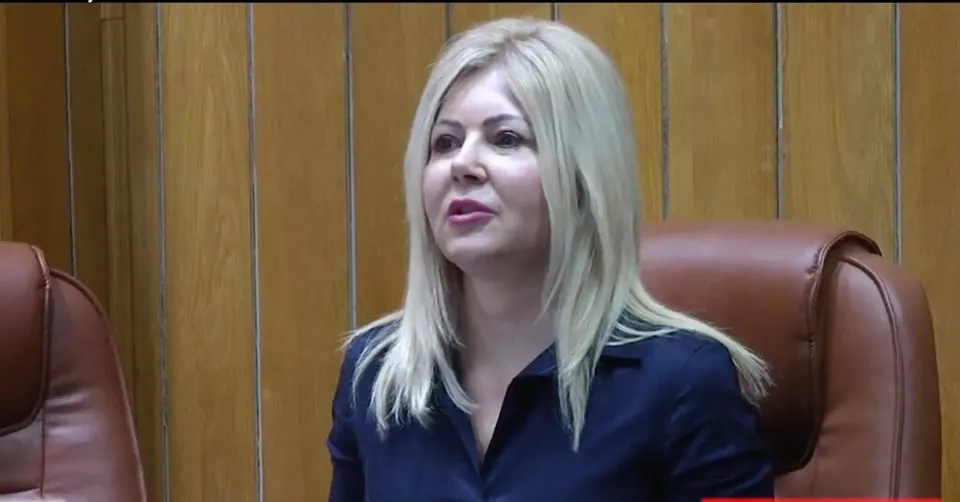 O femeie cu părul blond și o cămașă închisă la culoare stă în fața unui perete din panouri de lemn și a unor scaune de piele maro.