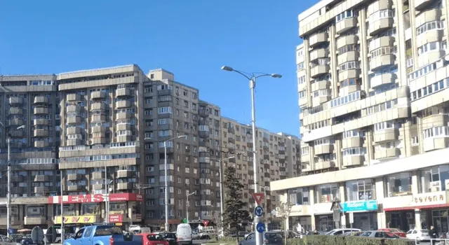 Blocuri înalte de apartamente cu balcoane de sticlă sunt situate pe o stradă cu mașini parcate și un stâlp de iluminat sub un cer senin.
