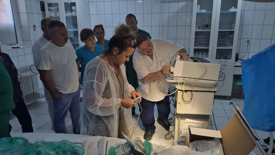 Un grup de persoane stă într-o încăpere cu plăci albe, privind un aparat medical alb aflat pe un suport metalic.