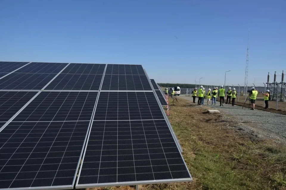 Rânduri de panouri fotovoltaice sunt instalate pe un teren lângă un grup de persoane purtând veste reflectorizante galbene sub un cer senin.