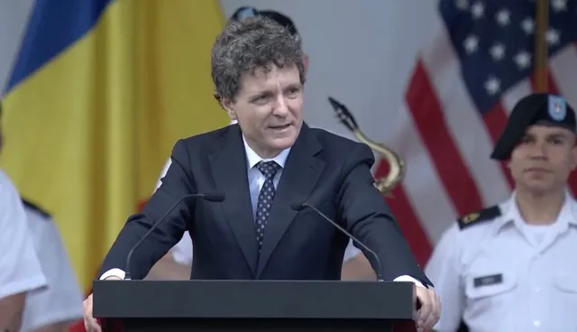 Un bărbat cu părul creț, îmbrăcat în costum închis și cravată, stă în spatele unui podium în fața unor steaguri.