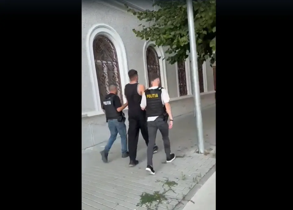 Trei bărbați, dintre care doi poartă veste negre cu inscripția POLITIA, merg pe un trotuar lângă o clădire cu ferestre arcuite.