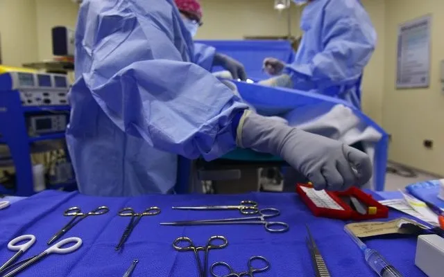 O persoană îmbrăcată în halat albastru și mănuși gri manipulează instrumente chirurgicale deasupra unui set de unelte metalice dispuse pe un material textil albastru.