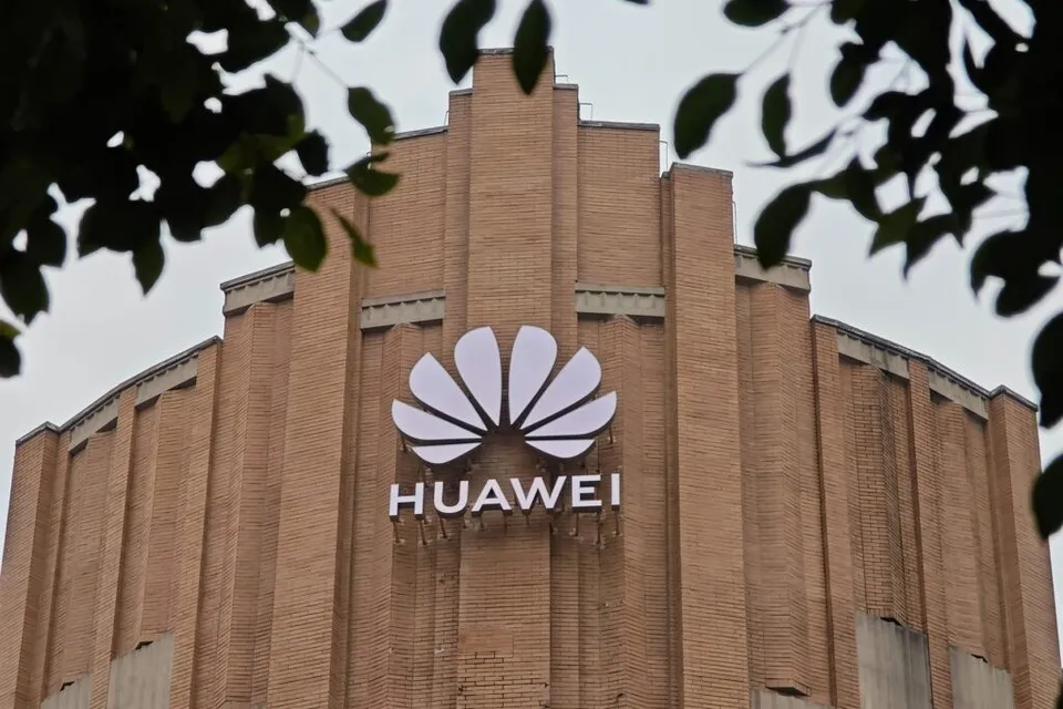 Un logo alb cu textul HUAWEI este montat pe fațada unei clădiri din cărămidă maro, încadrat de frunze verzi în prim-plan.
