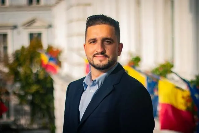Un bărbat cu barbă și păr scurt, purtând sacou închis și cămașă deschisă la guler, stă în fața unor clădiri și steaguri colorate.