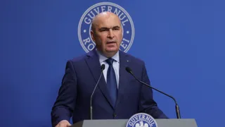Un bărbat cu costum închis și cravată stă în spatele unui podium cu microfoane în fața unui fundal albastru cu un sigiliu circular.