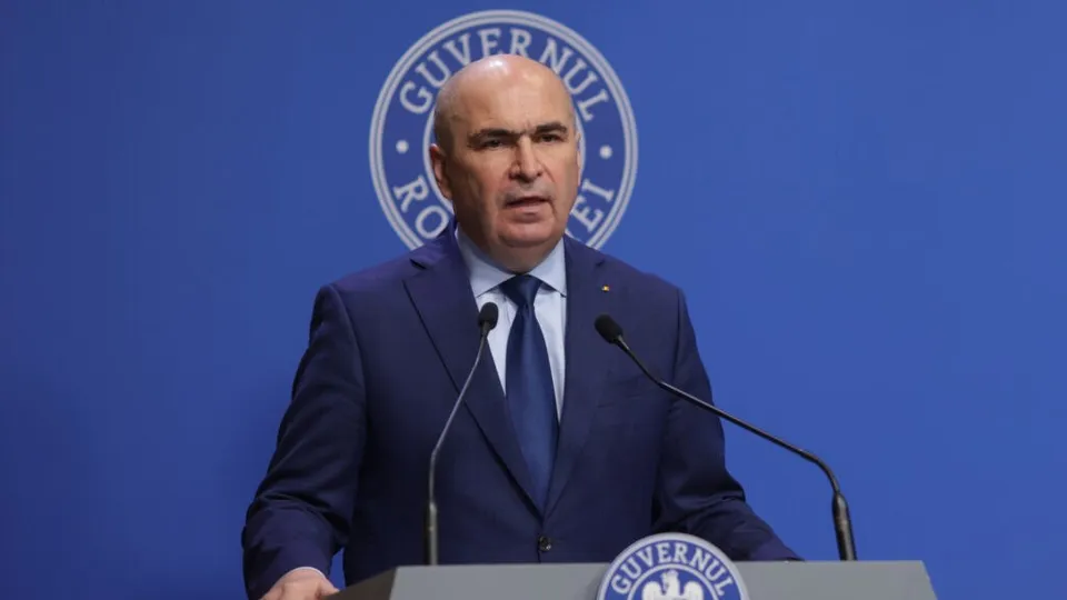 Un bărbat cu costum închis și cravată stă în spatele unui podium cu microfoane în fața unui fundal albastru cu un sigiliu circular.