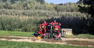 Un grup de persoane îmbrăcate în uniforme de protecție roșii și negre stau pe un teren verde lângă vegetație înaltă.
