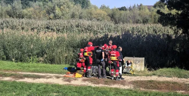 Un grup de persoane îmbrăcate în uniforme de protecție roșii și negre stau pe un teren verde lângă vegetație înaltă.