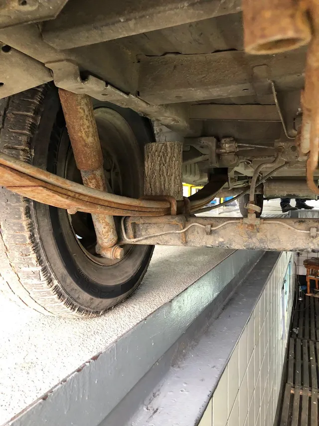 Partea inferioară a unui vehicul este vizibilă, afișând un roată, un amortizor ruginit și o piesă de lemn fixată la suspensie.