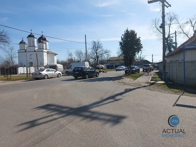 O stradă asfaltată cu mașini parate pe margini, o clădire albă cu turnuri și un stâlp de utilități sub un cer senin.
