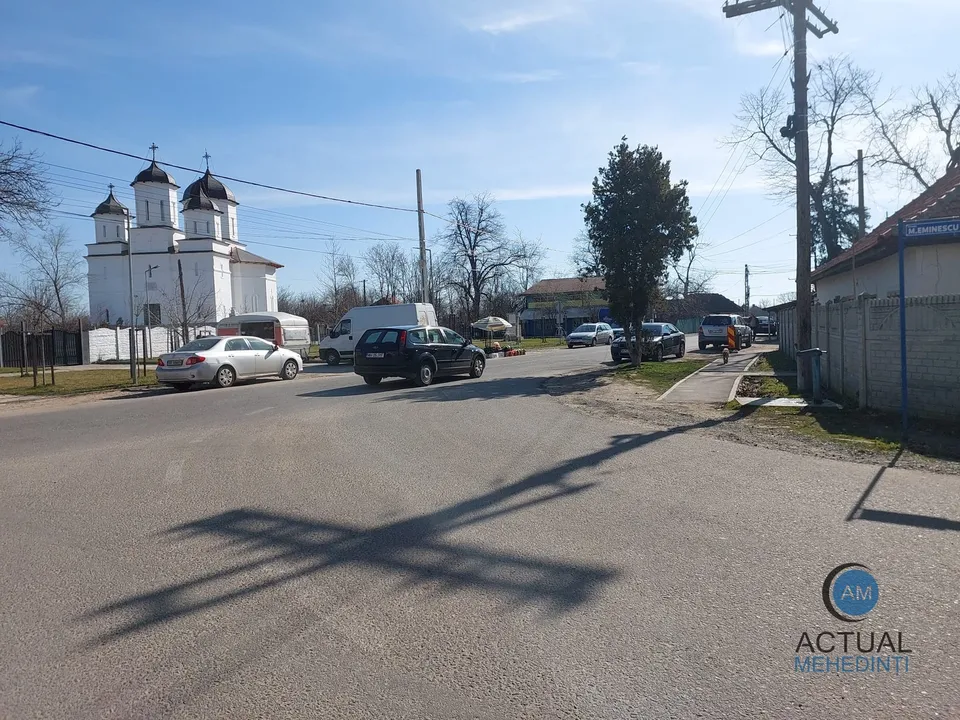 O stradă asfaltată cu mașini parate pe margini, o clădire albă cu turnuri și un stâlp de utilități sub un cer senin.