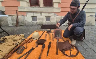 Un bărbat într-un uniformă albastră stă ghemuit lângă o masă acoperită cu obiecte metalice, piese de echipament și un rucsac pe o stradă pavată.