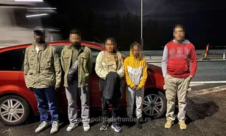 Patru persoane stau în picioare în fața unei mașini roșii pe un drum asfaltat, noaptea.