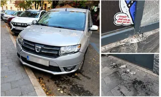 Un autoturism argintiu este parcat pe marginea unui trotuar lângă o clădire cu urme de deteriorări la baza geamului și pe pavaj.