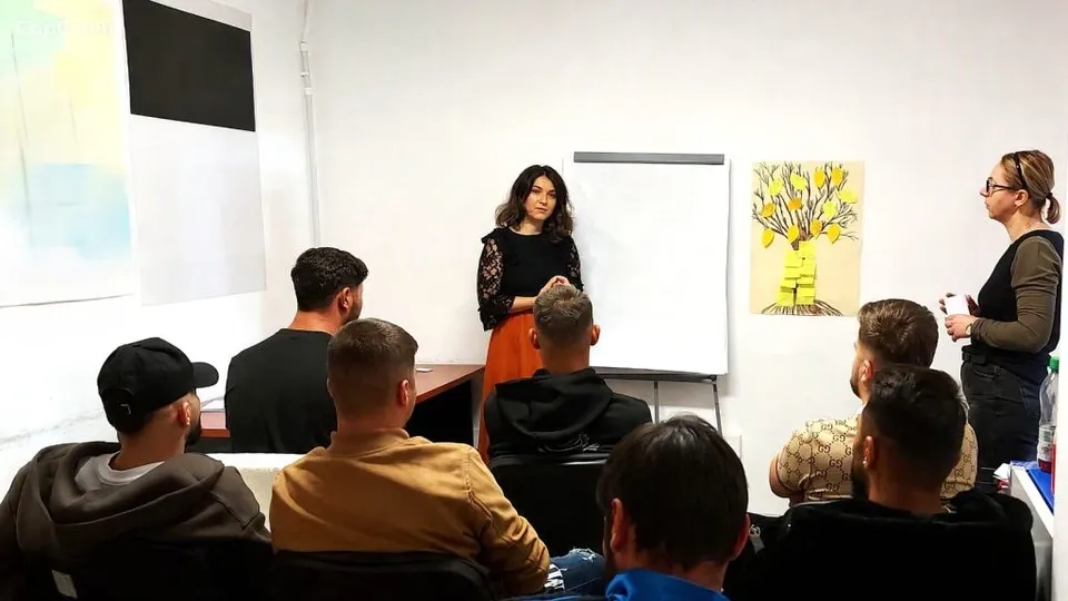 O femeie stă în picioare lângă un panou alb, în timp ce un grup de tineri stă pe scaune cu spatele la cameră.